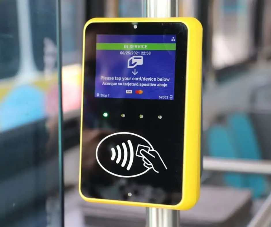 a yellow and black device is mounted on a pole just inside the bus' front door. It has a screed that says "in service, please tap your card/device below". There are Visa and Mastercard symbols on the screen. below the screen are four green lights, and below that is a white icon showing the contactless symbol: an oval with a wifi symbol on its side and a hand holding a square tag.