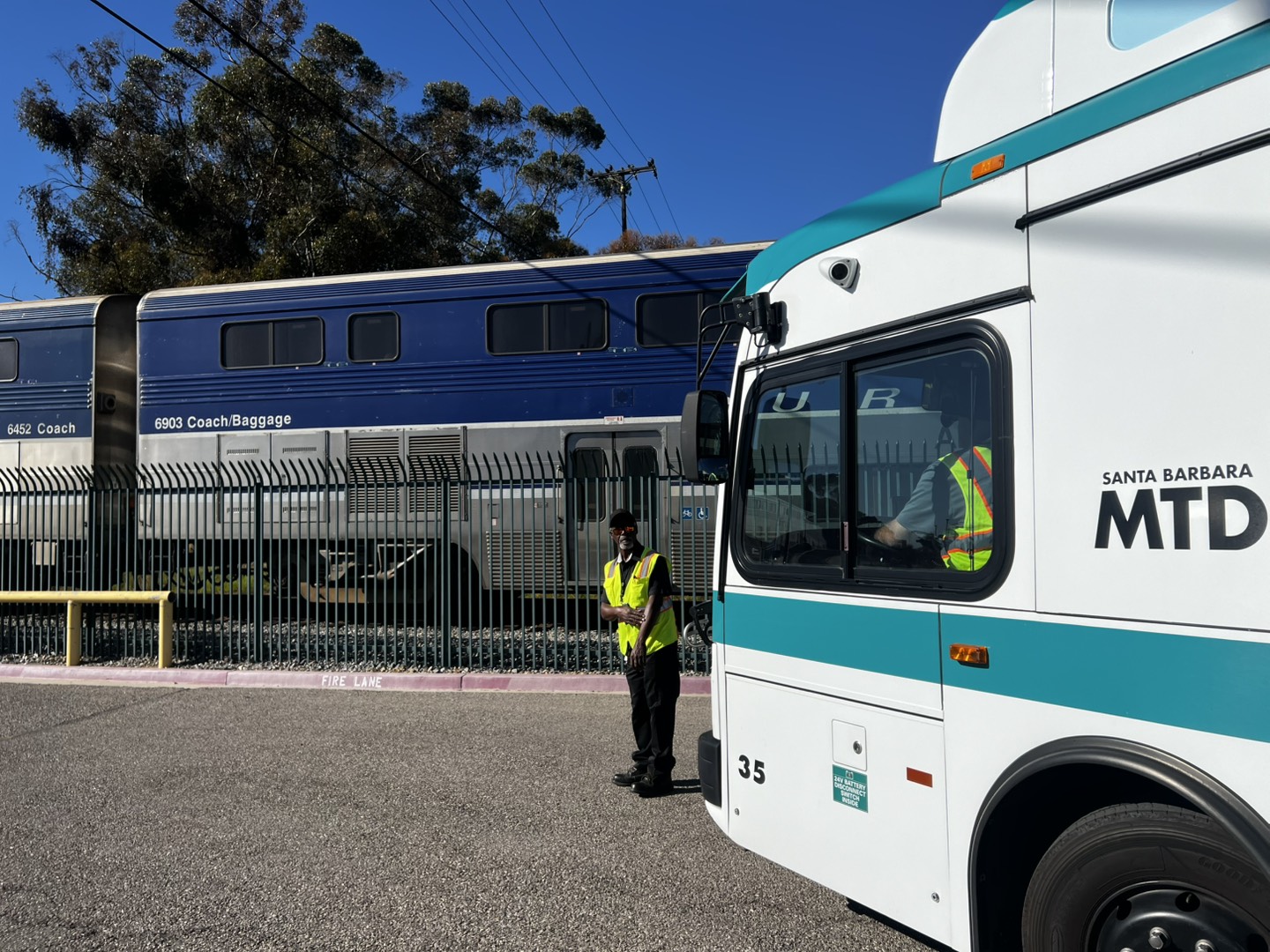 A photo of a First Mile Last Mile shuttle waiting for the Pacific Surfliner at the Amtrak station with a Bus Operator inside the shuttle and an Operations Supervisor standing outside the driver's side.