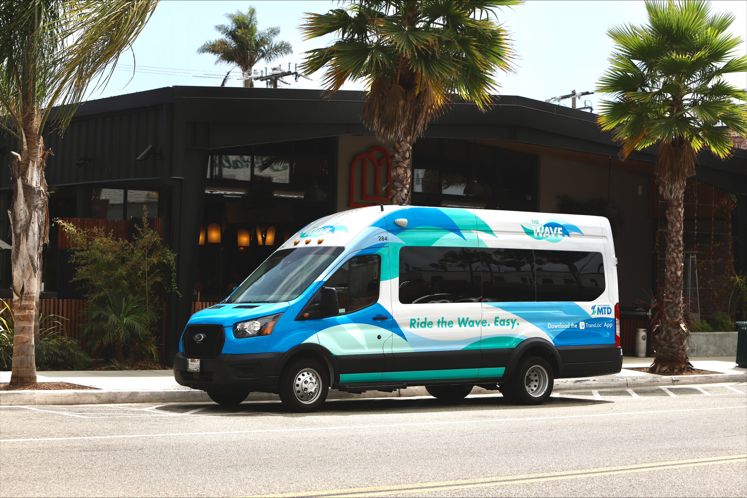 A photo of The Wave van in front of a black and brown building and palm trees in Carpinteria.
